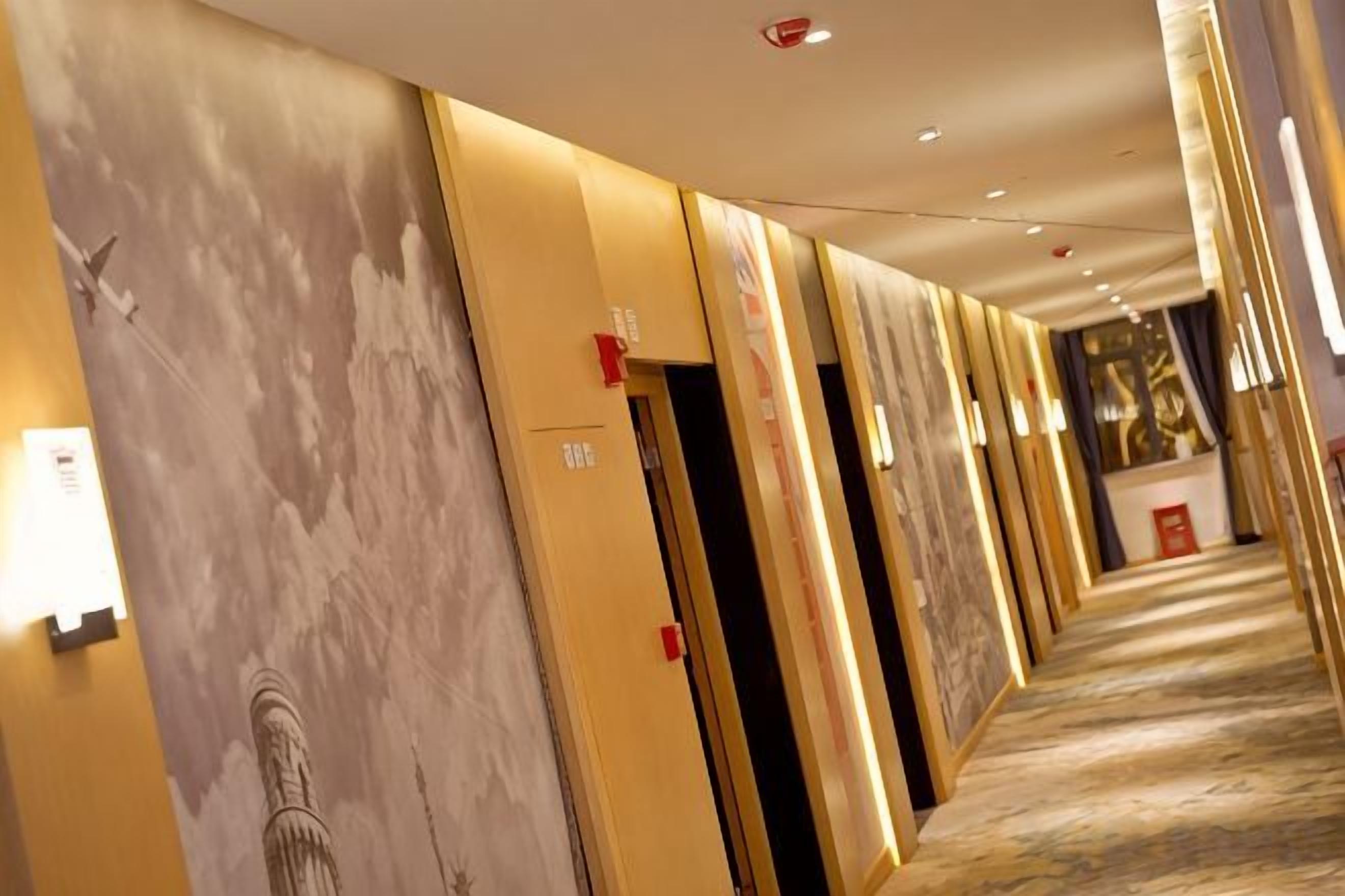 Un couloir d’hôtel avec des portes, un sol recouvert de moquette et un mur orné d’une fresque représentant un paysage urbain.