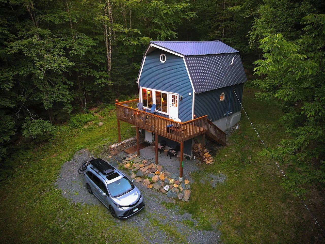 Una cabaña con terraza, un coche aparcado delante y un bosque circundante.