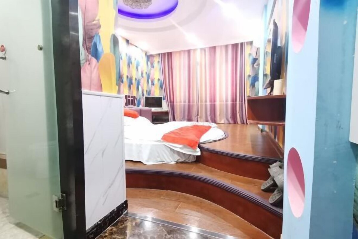 Habitación de hotel con una escalera curva, una cama con cubrecama blanco y rojo, un escritorio con una computadora y un mural abstracto colorido en la pared.