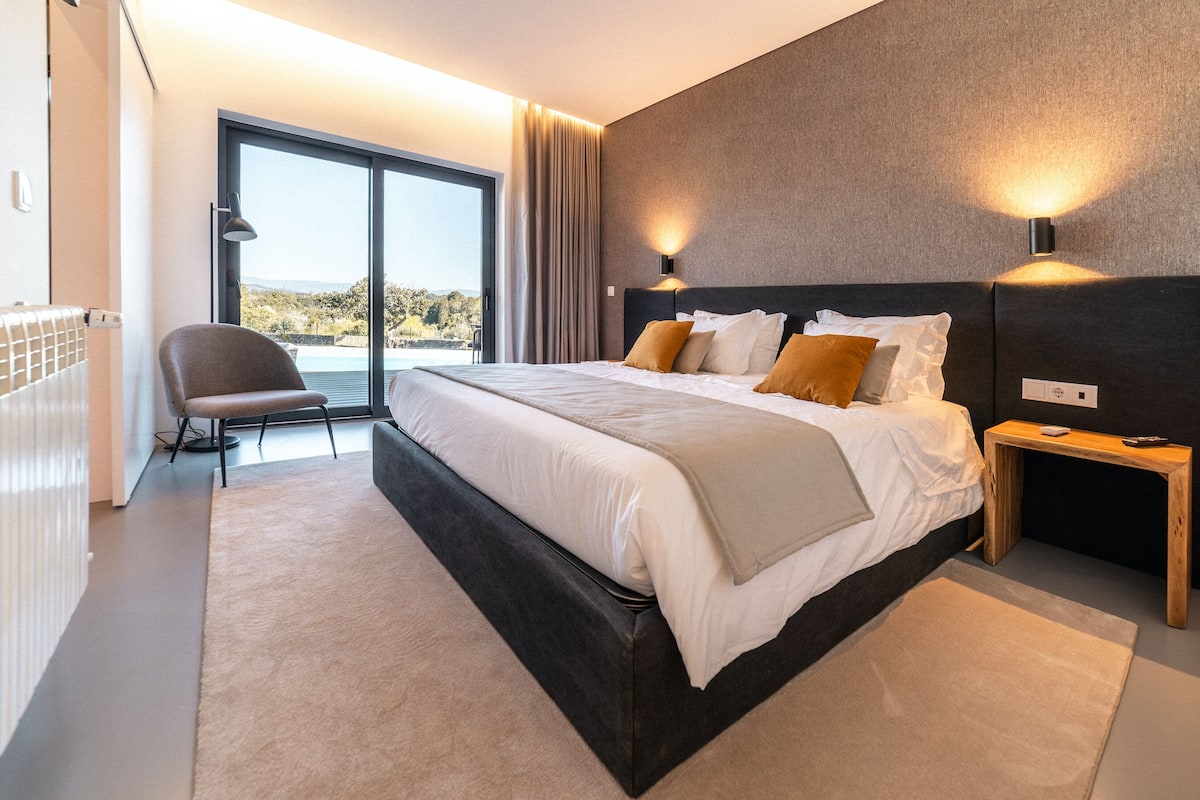A modern bedroom with a large bed, a chair, and a view of the outdoors.