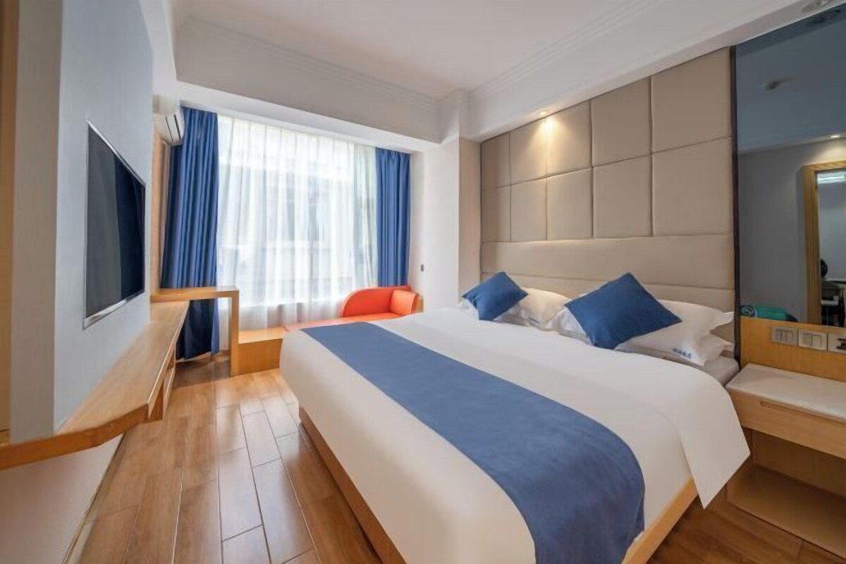A modern hotel room with a large bed, a TV, a seating area, and a window with curtains.