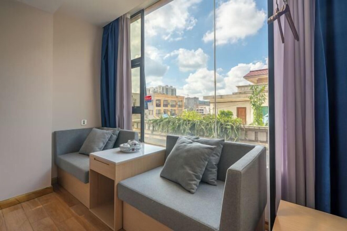 A modern room with a sofa and a small table, large windows with curtains, and a view of buildings and greenery outside.