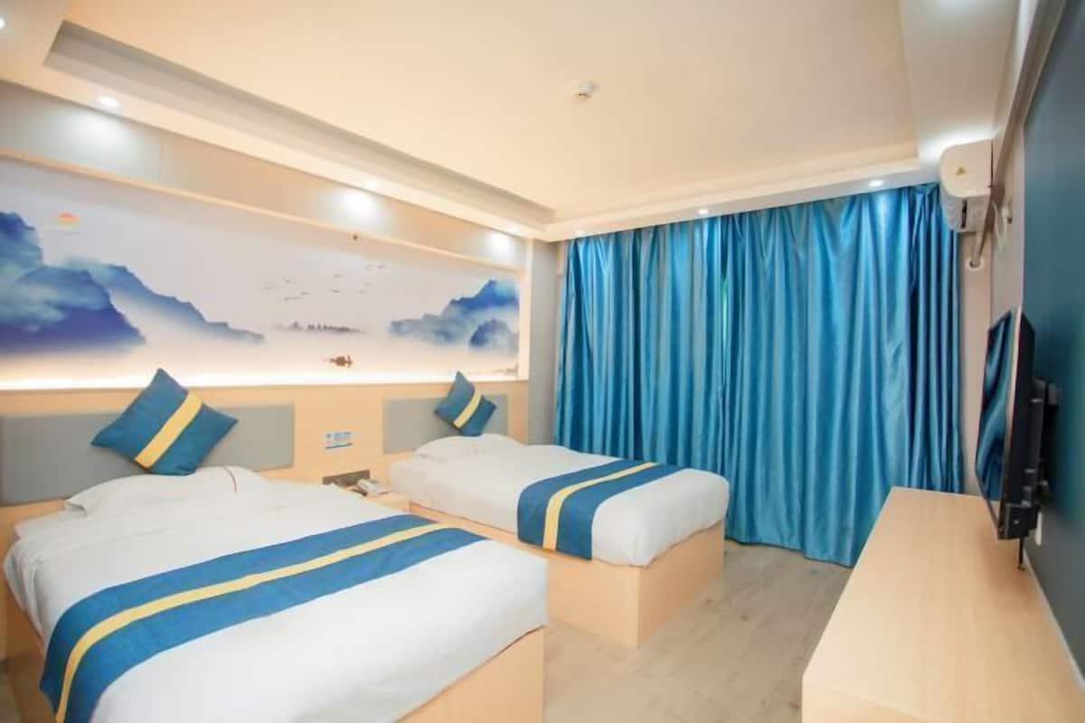 Habitación de hotel con dos camas, un escritorio, un televisor y un ventanal grande con cortinas.