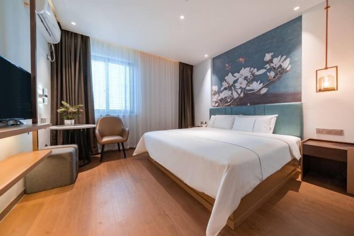 A hotel room with a large bed, a desk, a chair, a small table, and a wall-mounted TV.