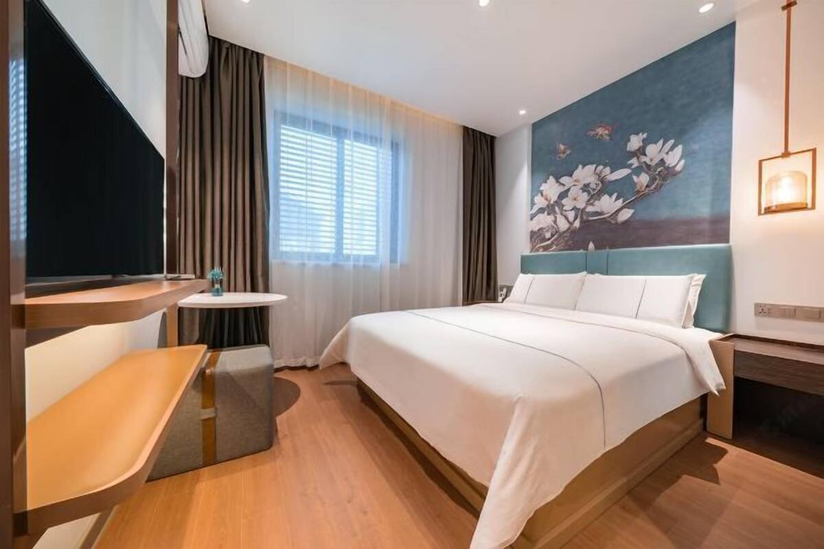 A hotel room with a bed, a desk, a small table, a window with curtains, and a wall-mounted light fixture.