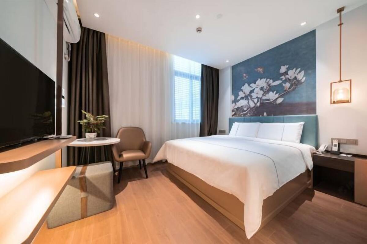 A modern hotel room with a large bed, a desk, a chair, a small table, a TV, and a painting on the wall.