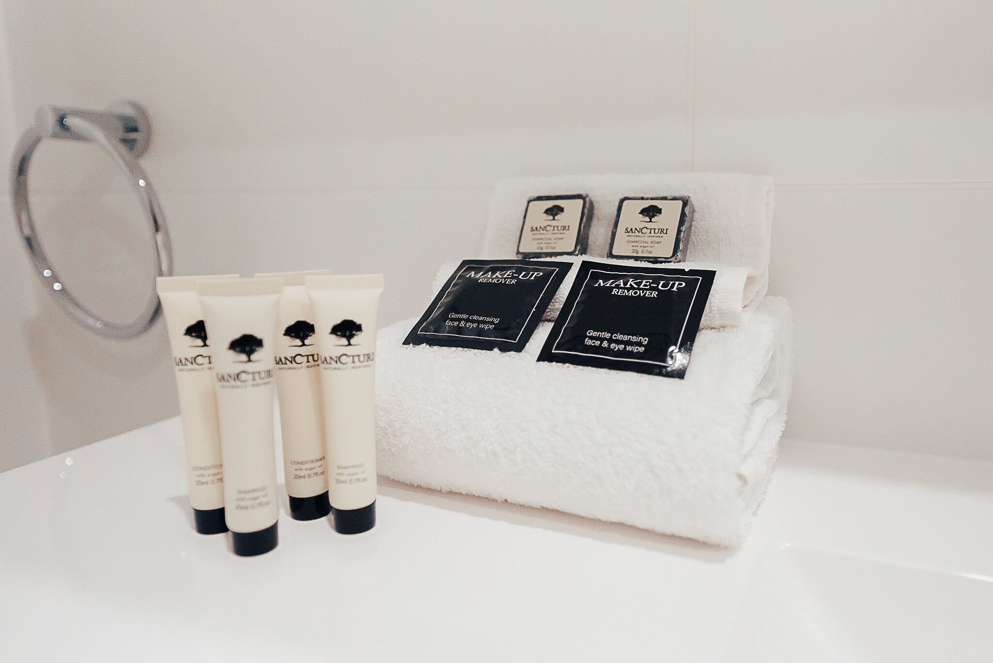 A bathroom with a white sink, a white towel, and three bottles of 'Sanctuary' makeup remover.