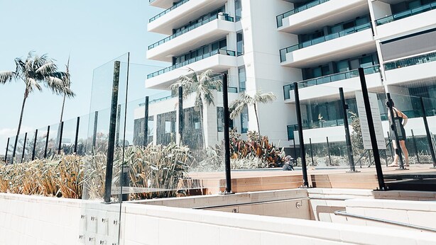 Un edificio moderno de varios pisos con balcones, un paseo cubierto y palmeras.