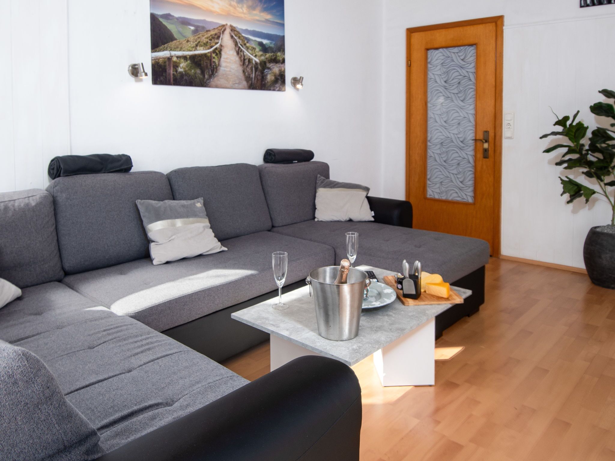 A modern living room with a grey sectional sofa, a small coffee table with a champagne bottle, glasses, and a cheese board.