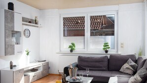 Ein modernes Wohnzimmer mit einem grauen Ecksofa, einem kleinen Esstisch mit Schneidebrett und Käse, einer Küchenzeile mit grauem Schrank und einer Uhr sowie einem Fenster mit Jalousien und Topfpflanzen.