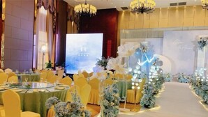 A banquet hall with round tables, yellow chairs, floral centerpieces, and a stage with a backdrop. - Lijing Hotel (Ningbo)