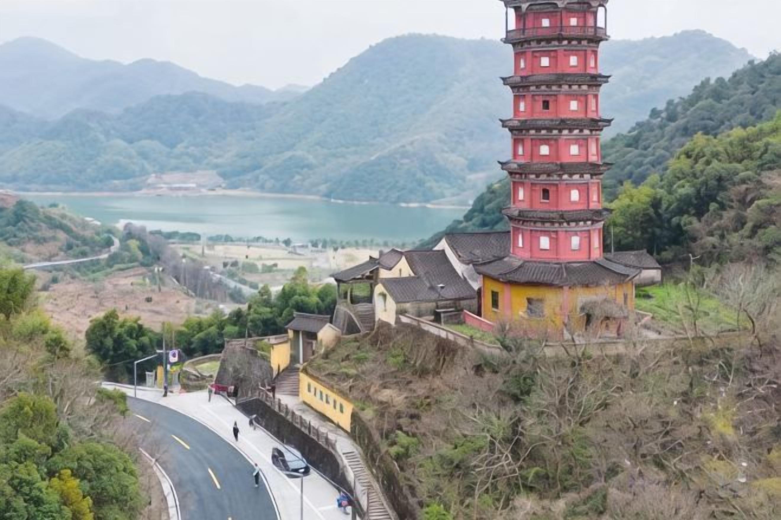A pagoda-style tower with a winding road leading to it, surrounded by greenery and a body of water in the background.