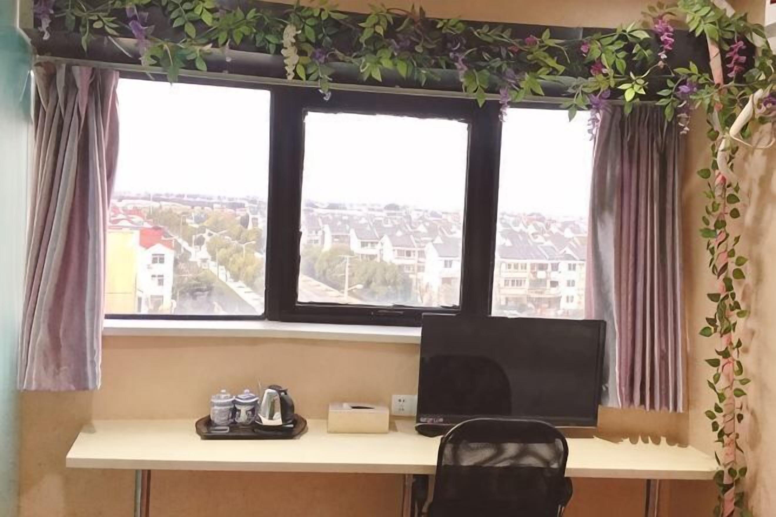 A room with a desk, a laptop, a window with curtains, and a view of buildings.