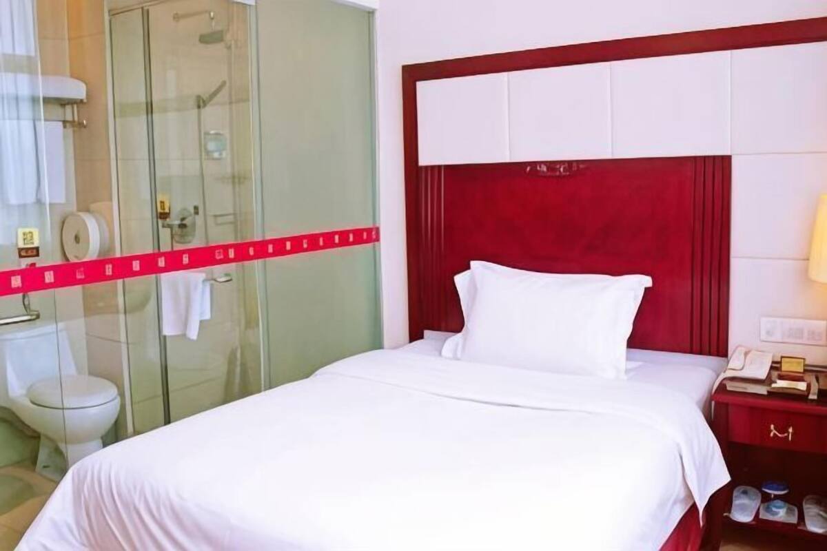 A hotel room with a bed, a red headboard, a glass shower enclosure, a white toilet, and a red nightstand with a lamp.