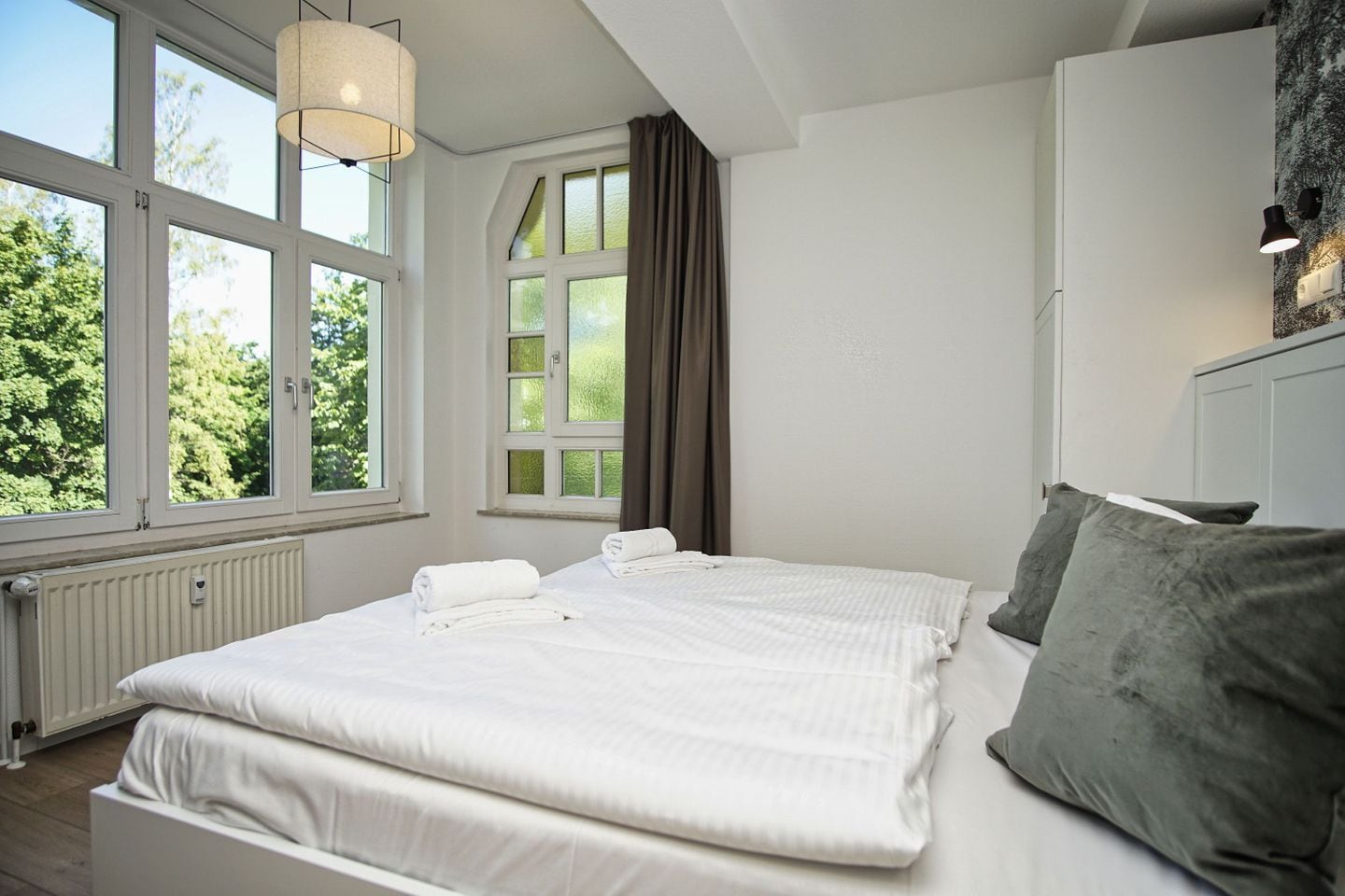 Ein Schlafzimmer mit einem großen Bett, einem Fenster mit Vorhängen, einem Heizkörper und einer Wandleuchte.