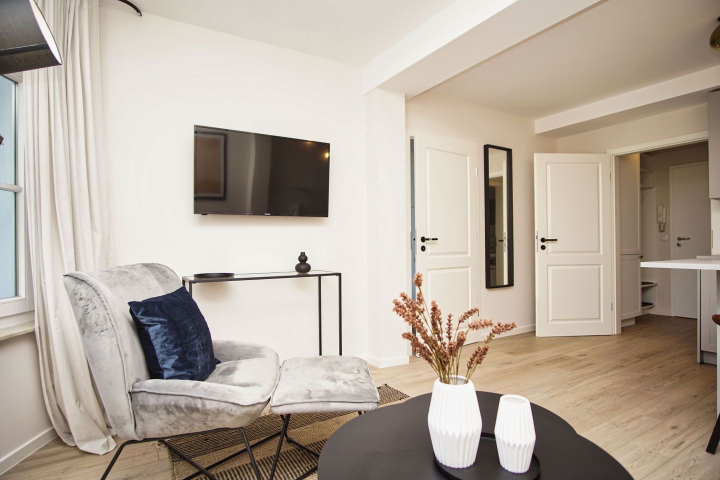 Un salon moderne avec un fauteuil gris, un téléviseur à écran plat, une petite table ornée de vases décoratifs et une porte-fenêtre donnant sur une cuisine.