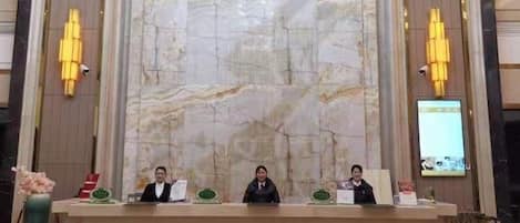 Un mostrador de recepción con tres personas sentadas detrás, una pared de mármol al fondo y dos lámparas fijadas a la pared.