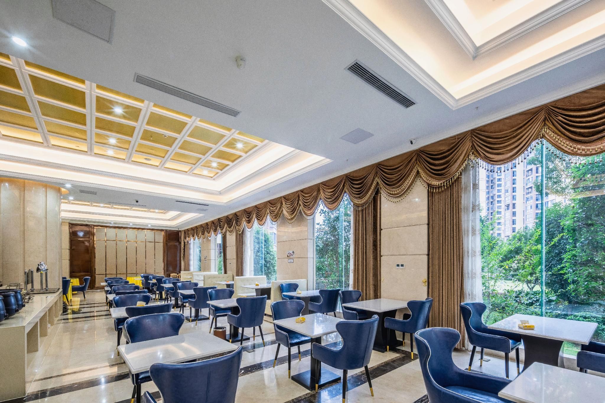 A hotel dining area with tables and chairs, large windows with curtains, and a ceiling with skylights.