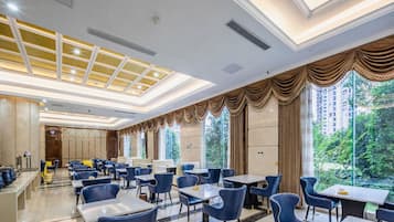 A hotel dining area with tables and chairs, large windows with curtains, and a ceiling with skylights.