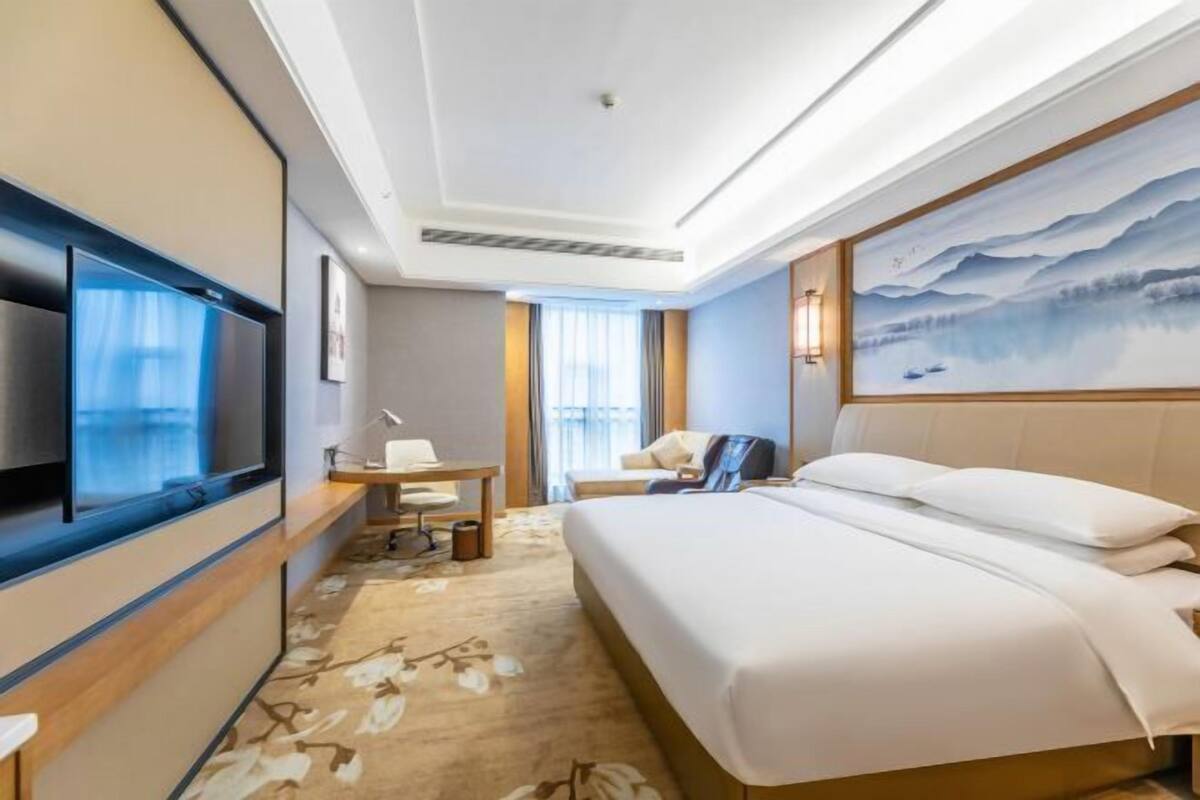 A hotel room with a bed, a TV, a desk, and a painting on the wall.