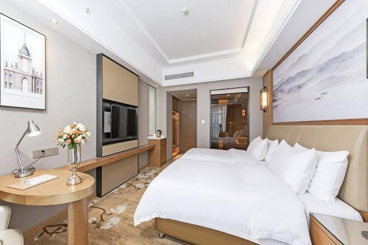 A hotel room with a bed, a desk with a lamp, a TV, a painting, and a view of a building.