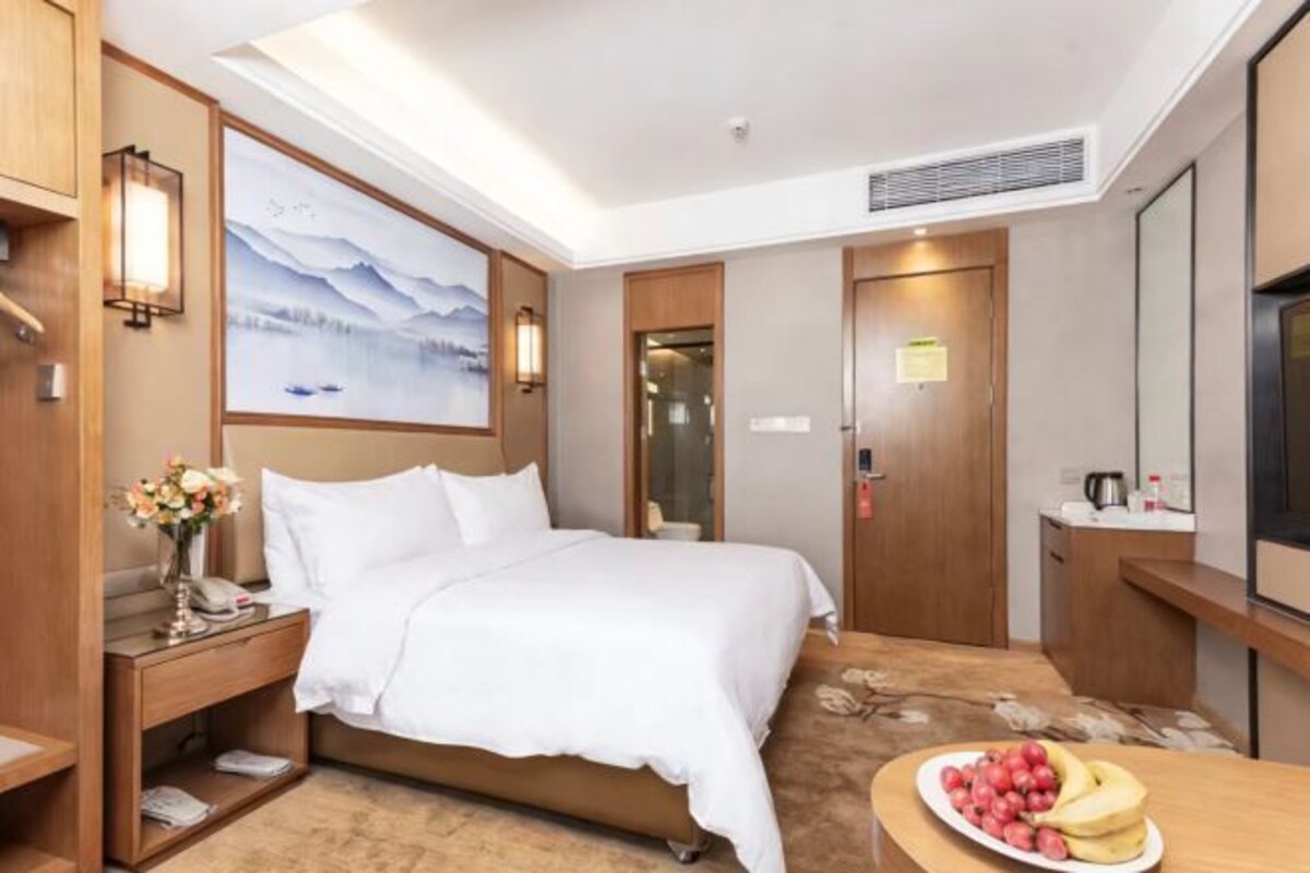 A hotel room with a bed, bedside table, lamp, artwork, and a fruit bowl.