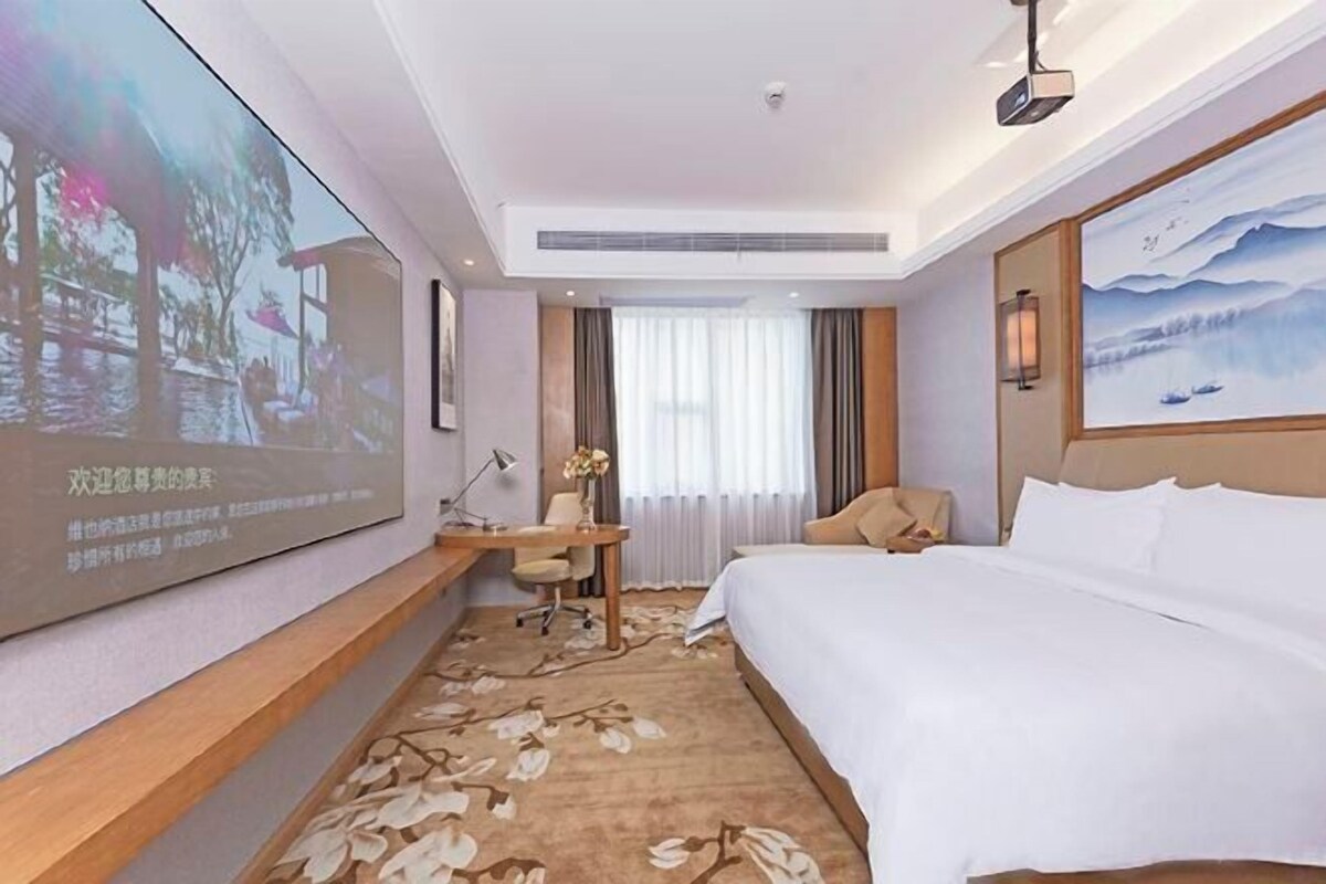 A hotel room with a bed, desk, chair, and a large screen displaying a scenic image.