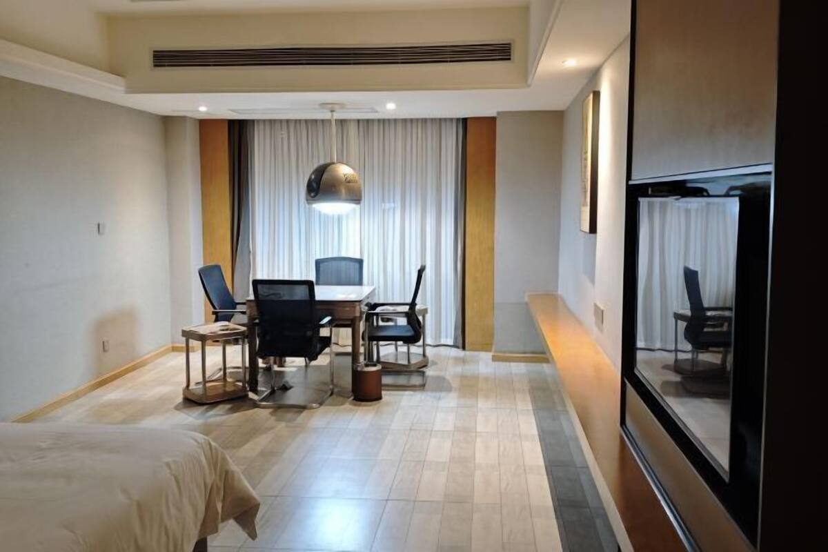 A hotel room with a bed, a desk with a computer, two chairs, a large window with curtains, and a mirror on the wall.
