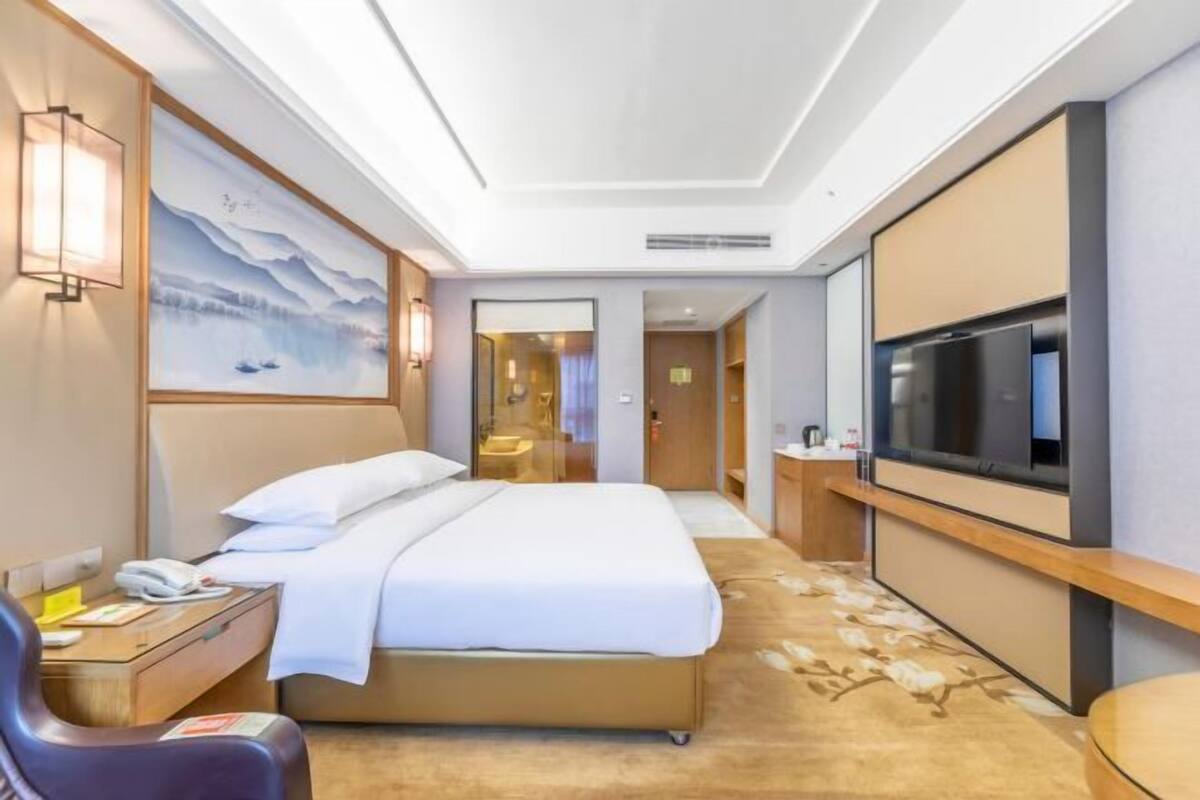 A hotel room with a bed, bedside table, lamp, TV, and a painting on the wall.
