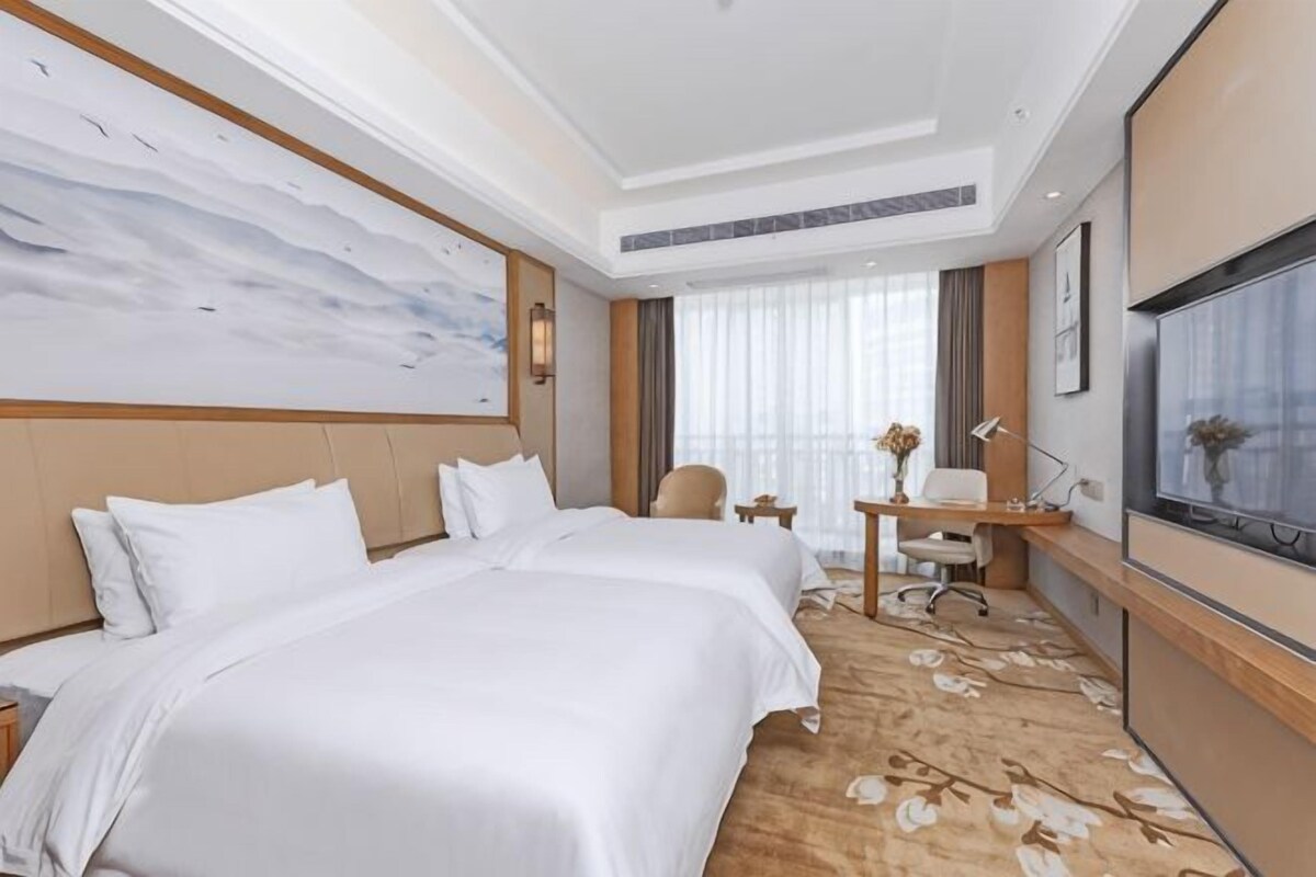 A hotel room with two beds, a desk, a chair, a TV, and a large window.