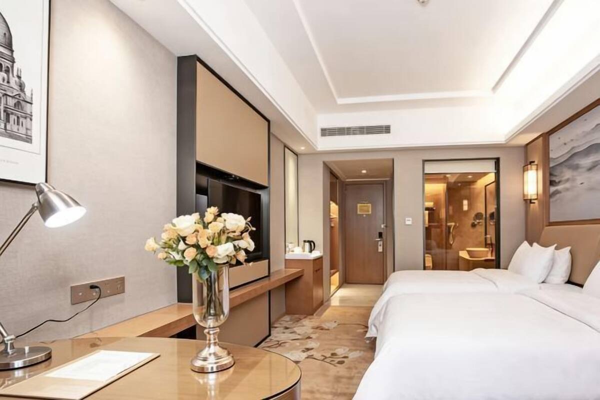 A hotel room with a bed, bedside table, lamp, TV, and a vase with flowers.