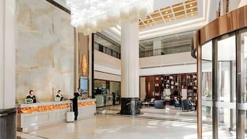 A spacious lobby with a marble reception desk, a large chandelier, and a multi-level atrium with a glass railing.