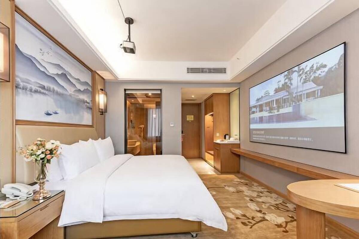 A hotel room with a bed, a desk with a phone, a vase with flowers, a large painting, a projection screen, and a carpet.