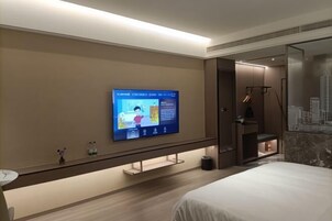Quarto de hotel moderno com uma grande televisão de tela plana na parede, cama com roupa de cama branca, piso de madeira e uma pequena mesa com um vaso.