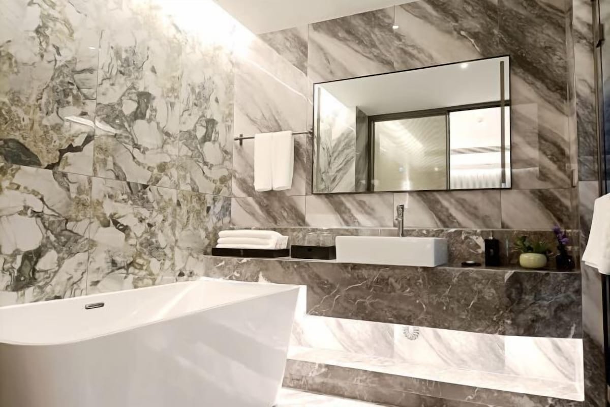 A modern bathroom with a freestanding bathtub, a large mirror, and marble walls.