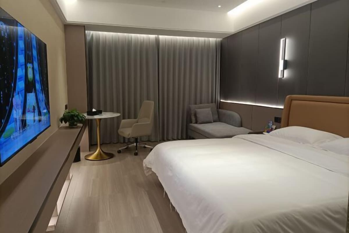 A modern hotel room with a large bed, a desk with a chair, a flat-screen TV, and a large window with curtains.