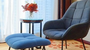 A modern room with a blue armchair and ottoman, a glass table with a vase of flowers, and a view of the city through sheer curtains. - The BLD International Hotel (Yuncheng)
