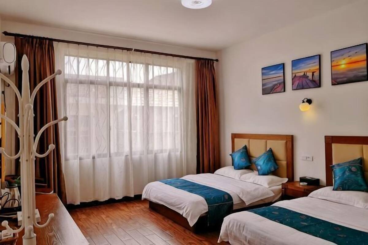 A hotel room with two beds, a large window with curtains, a coat rack, and wall art.