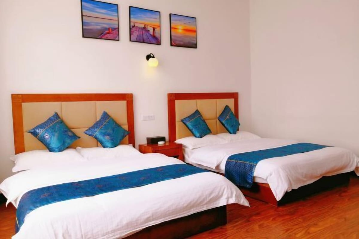 A hotel room with two beds, wooden headboards, white bedding, blue decorative pillows, a nightstand with a lamp, and three framed pictures on the wall.