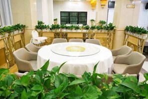 Ein runder Tisch mit weißer Tischdecke, umgeben von beigen Stühlen, in einem Raum mit goldener Geländer und Topfpflanzen.