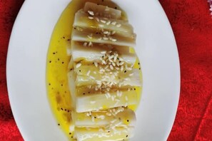 Un plato blanco con pescado blanco en rodajas, adornado con semillas de sésamo y pimienta negra, sobre un mantel rojo.