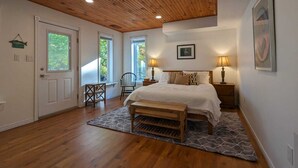 Un dormitorio con techo de madera, una cama con cubrecama blanco, dos lámparas de noche, una silla y una mesita.