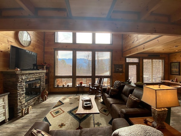 L’intérieur chaleureux d’un chalet avec une cheminée en pierre, un canapé en cuir, une table à manger et de grandes fenêtres donnant sur le paysage extérieur.