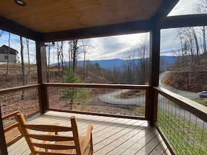 Une terrasse en bois avec un banc, une vue sur une route sinueuse et une chaîne de montagnes à l’horizon.