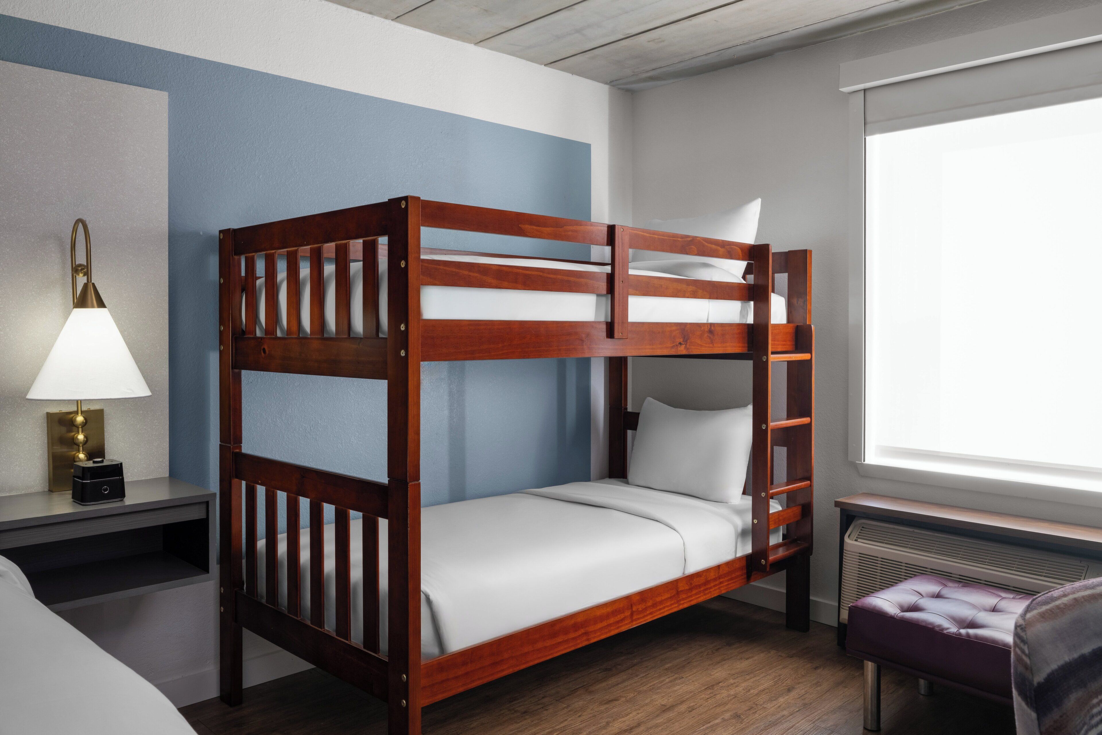 A bunk bed with a wooden frame, a bedside table with a lamp, and a window with a view.