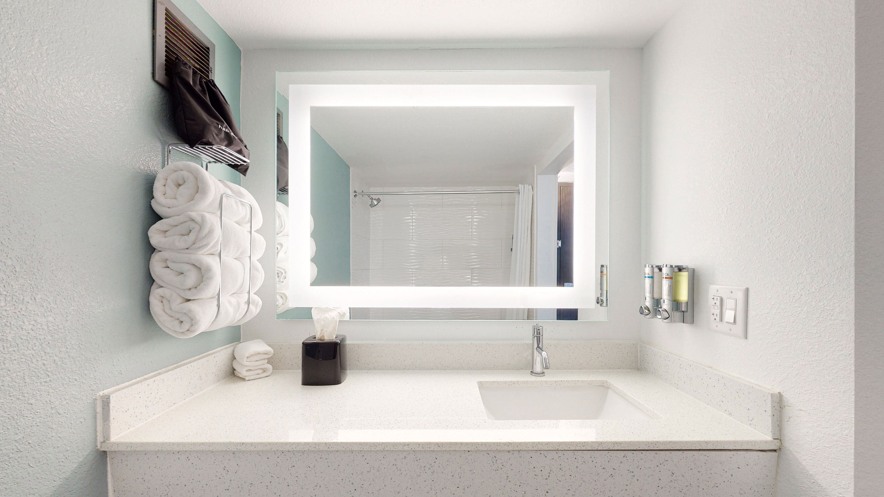 A bathroom with a white sink, a towel rack with folded towels, a tissue dispenser, and a mirror.
