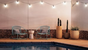Zona de piscina con dos sillas color turquesa, una mesa blanca y dos grandes cactus en macetas, todo frente a un muro de ladrillo.