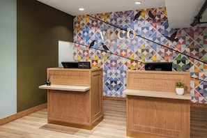 Un espace d’accueil doté de deux comptoirs de réception en bois, d’un mur décoré de motifs géométriques colorés et d’un plafond où les tuyaux sont apparents.