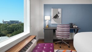 Habitación de hotel con una ventana grande, un escritorio con silla, una cama y vista a la ciudad.
