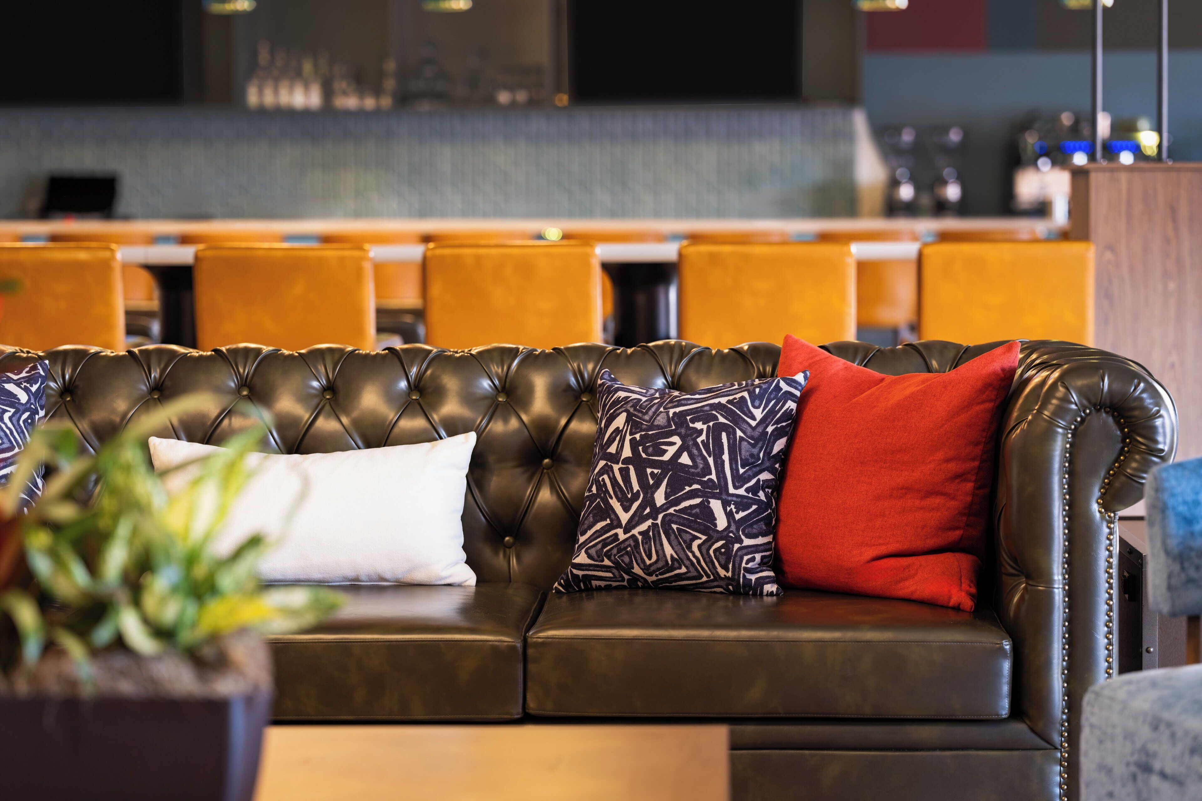 A leather sofa with patterned and solid pillows, a potted plant, and a bar area in the background.